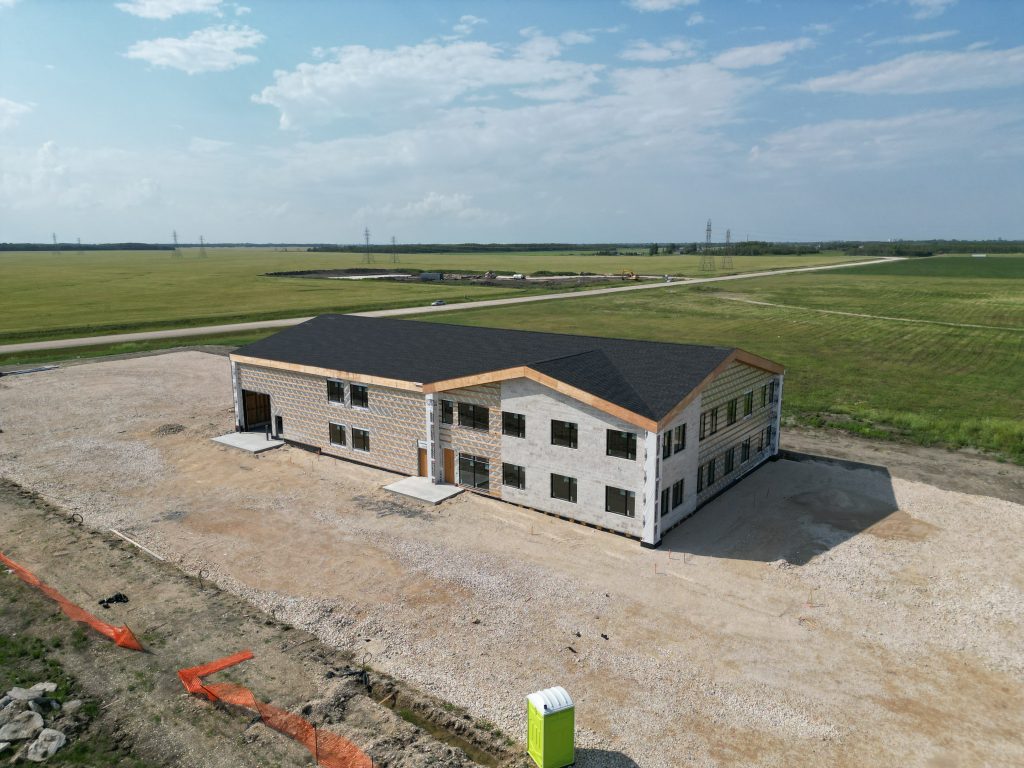Two-story unfinished building with a black roof on a dirt lot in a wide open field; construction equipment in distance.
