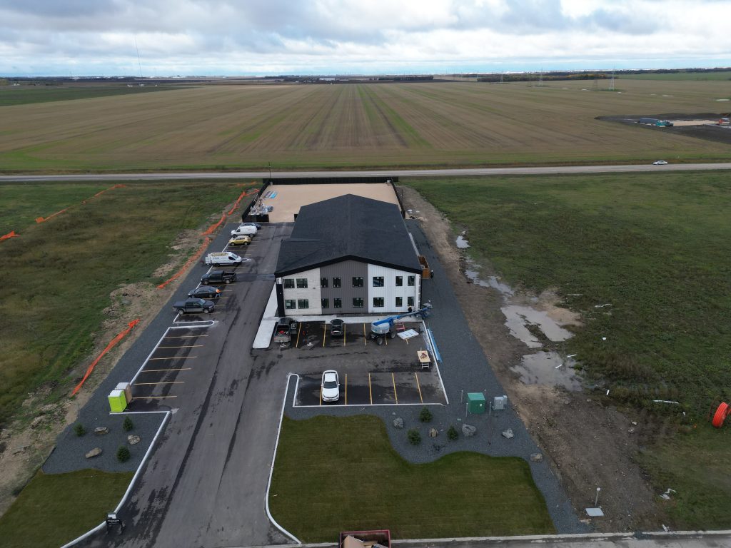 Aerial view of a white two‑story office building with a dark roof, a surrounding parking lot with several cars, and open farmland beyond it.